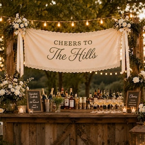 May include: A cream-colored fabric banner with lace trim featuring the text "CHEERS TO The Hills" in dark lettering, suspended between two wooden posts decorated with white floral arrangements and string lights above a wooden bar.