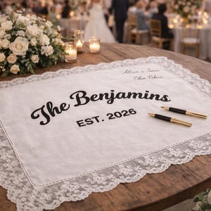 May include: A white, lace-edged wedding guest book with the words "The Benjamins EST. 2026" in black script. Two gold and black pens rest on the book, ready for signatures. The scene is set at a wedding reception.