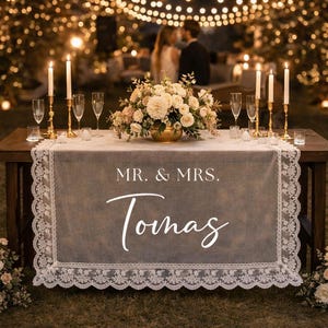 Könnte beinhalten: Ein dekorierter Tisch für eine Hochzeitsfeier, mit einem grauen Tischläufer mit weißer Spitzenborte. Der Läufer zeigt den Text "MR. & MRS. Tomas" in weißer Schrift. Der Tisch ist mit Blumen, Kerzen und Sektgläsern geschmückt.