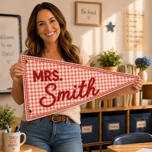 Puede incluir: Un banderín triangular de tela con un patrón de cuadros vichy rojos y blancos y un borde de pompones blancos. El texto MRS. Smith está bordado en letras cursivas rojas grandes y en relieve en el centro. Tiene dos ojales de latón arriba.