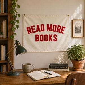 May include: A rectangular off-white fabric wall hanging featuring the red text READ MORE BOOKS in a bold, arched font. The banner includes metal grommets in the top corners for mounting and is displayed on a wall above a wooden desk.
