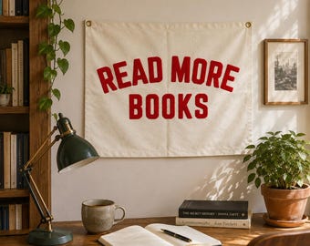 May include: A rectangular off-white fabric wall hanging featuring the red text READ MORE BOOKS in a bold, arched font. The banner includes metal grommets in the top corners for mounting and is displayed on a wall above a wooden desk.