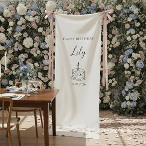 May include: A white fabric banner with the text "HAPPY BIRTHDAY Lily" and a cake illustration. The banner is suspended with pink ribbons against a backdrop of flowers. A wooden table is set for a meal.