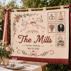 Könnte beinhalten: Ein cremefarbenes Hochzeitsbanner mit gestickten Details. Es zeigt die Worte „Just Married“ und „The Mills“ sowie das Datum 24. Mai 2026. Das Banner ist mit Tauben, Ringen, einer Torte, einem Smoking und einem Auto mit Sektgläsern verziert.