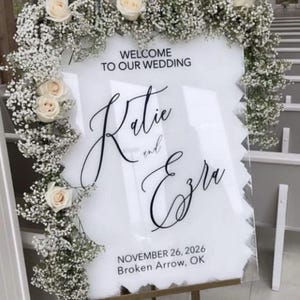 May include: A white wedding welcome sign with the names "Katie and Ezra" in elegant script. The sign is framed with white roses and baby's breath. The date and location are also displayed.
