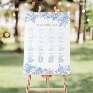May include: A white wedding seating chart sign on a wooden easel featuring blue floral illustrations at the top and bottom borders. The text reads FIND YOUR SEAT followed by a list of names organized into twelve tables in a blue serif font.