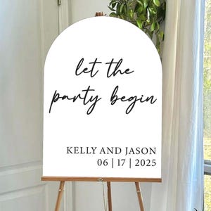 May include: White arched sign on a wooden easel with the text "let the party begin" in black script. Below, it reads "KELLY AND JASON 06 | 17 | 2025". The sign is displayed indoors near a window.