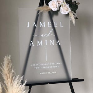 May include: A frosted acrylic wedding sign with the names "JAMEEL and AMINA" in white script font. The sign also includes the date "MARCH 23, 2024" and the text "ARE DELIGHTED TO WELCOME YOU TO THEIR WEDDING".