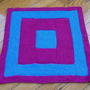 May include: A knitted square throw blanket with a concentric square pattern. The outer border is blue, the next layer is pink, the next layer is blue, and the center square is pink.