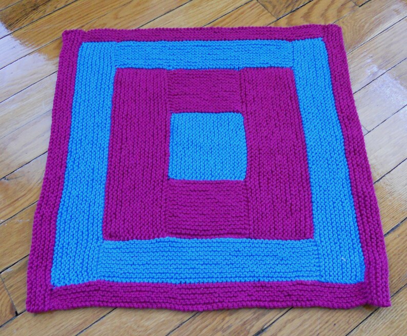 May include: A knitted square throw blanket with a concentric square pattern. The outer border is blue, the next layer is pink, the next layer is blue, and the center square is pink.