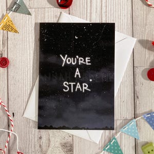 May include: A greeting card with the words "You're A Star" set against a dark, starry sky. The card is in a white envelope, with bunting and buttons arranged on a wooden surface.
