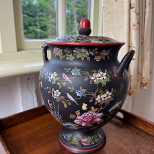 Puede incluir: Un jarrón decorativo de cerámica negra con tapa, con diseños florales y de pájaros en rosa, azul, amarillo y verde. El jarrón tiene un borde rojo y un adorno rojo y negro en la tapa. Se encuentra sobre una bandeja de madera.
