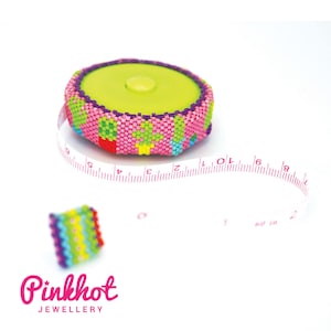 May include: A pink and green beaded tape measure with a white measuring tape extending from it. The tape measure is decorated with colorful beads and has a small, colorful beaded ring next to it. The text "Pinkhot Jewellery" is visible at the bottom of the image.