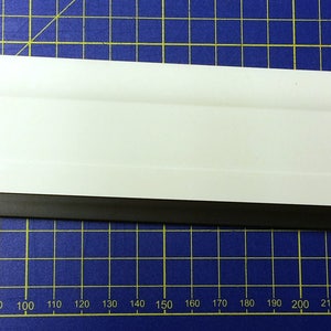 May include: A white and black squeegee with a rubber blade. The squeegee is on a blue gridded surface.