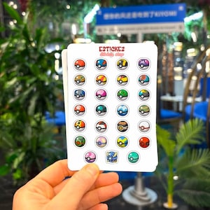 May include: A collection of stickers displaying diverse, colourful Poke Ball designs. The stickers are presented on a white card, held by a hand. The text "ESTICKEZ" is visible at the top of the stickers.