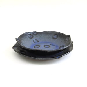 May include: Two-piece ceramic serving set. The set includes a small, dark blue bowl with a wavy rim and a matching plate. The bowl has a blue interior with dark blue circles. The plate is dark blue with a slightly raised edge. The set is on a white background.
