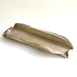 May include: A long, beige ceramic tray with a leaf-like shape. The tray has a textured surface with parallel lines running along its length. The edges are slightly raised and have a darker brown color.