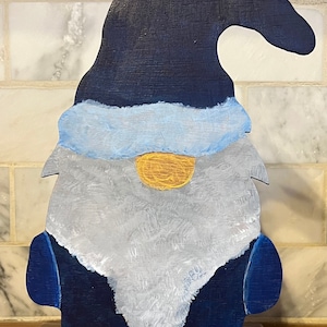 Puede incluir: Una figura de gnomo de madera pintada en tonos de azul, blanco y amarillo. El gnomo tiene un sombrero largo y puntiagudo, una barba blanca y una nariz amarilla. El gnomo está de pie sobre una base de madera.