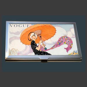 May include: A vintage Vogue magazine cover featuring a woman in a black dress holding an orange parasol with a pink and purple scarf blowing in the wind.