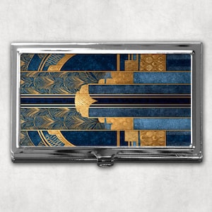 Peut inclure: Un étui à cartes de visite en métal argenté avec un motif géométrique. Le motif présente des motifs dorés et bleu marine, notamment des feuilles stylisées et des rayures horizontales. L'étui a une ouverture à charnière.