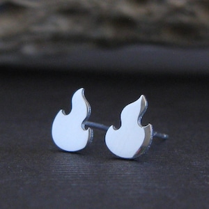 Puede incluir: Un par de pendientes de botón plateados con forma de llamas. Los pendientes son pequeños y tienen un acabado pulido. El fondo es una superficie gris oscuro con una textura de madera borrosa.