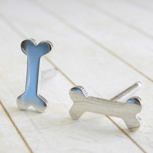 Puede incluir: Un par de pendientes de plata en forma de hueso. Los pendientes son pequeños y delicados, y serían un gran regalo para un amante de los perros.