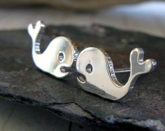 May include: A pair of silver whale-shaped stud earrings. Each earring features a stylized whale design with a curved body, a small eye, and a raised tail fin. The earrings are displayed on a dark, textured surface.