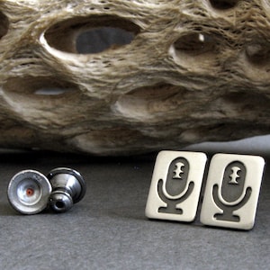 Peut inclure: Une paire de boutons de manchette en argent avec un motif de microphone. Les boutons de manchette sont rectangulaires et ont une surface légèrement texturée.