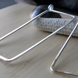 May include: A pair of silver rectangular hoop earrings. The earrings are made of a thick, polished metal and have a simple, modern design.