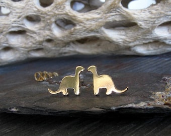 May include: A pair of gold dinosaur earrings. The dinosaurs are facing each other and are in a simple silhouette style.