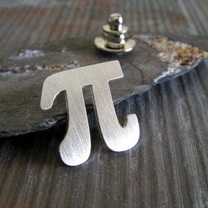 May include: Silver-toned metal pin shaped like the mathematical symbol pi. The pin has a brushed finish and a simple design.