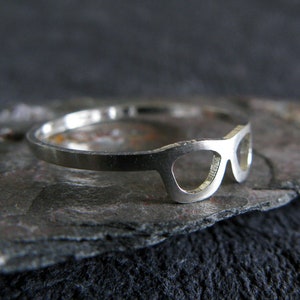 May include: A silver ring with a cut-out design of sunglasses. The ring is on a dark gray background.