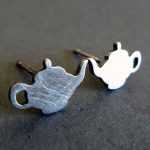 May include: A pair of silver teapot-shaped stud earrings. The earrings are made of metal and have a brushed finish.