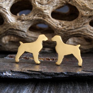 May include: A pair of gold-colored dog-shaped stud earrings. The earrings are in the shape of a dog, with a detailed outline. The earrings are displayed on a dark gray surface, with a textured wooden background.