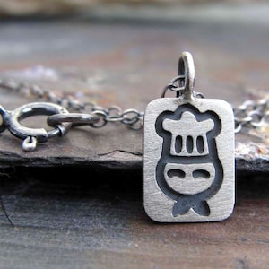 Könnte beinhalten: Eine silberne Halskette mit einem rechteckigen Anhänger, der ein Kochmützen- und Gesichtsdesign zeigt.