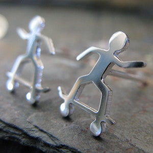 May include: Two silver stick figure earrings, each depicting a person skateboarding. The earrings are detailed and have a modern, minimalist design.