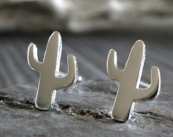 May include: A pair of silver-toned stud earrings shaped like saguaro cacti. Each earring features a smooth, polished metallic surface with a central vertical column and two smaller side arms extending outward, designed as simple silhouettes.