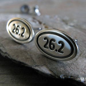 Puede incluir: Un par de pendientes ovalados de plata con el número "26.2" grabado en cada pendiente.