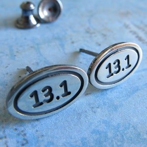 May include: A pair of silver earrings with a rounded oval shape. Each earring features the number "13.1" engraved in black.