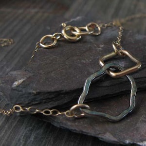 May include: A delicate gold-toned necklace with a unique design. The necklace features a chain and a pendant with a geometric shape and a textured, irregular shape. The necklace is displayed on a dark, textured surface.