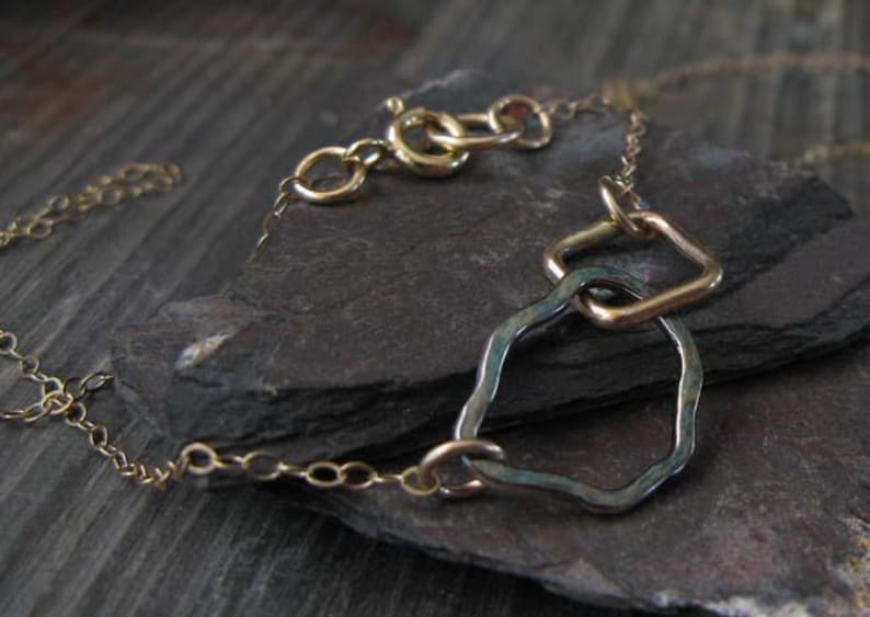 May include: A delicate gold-toned necklace with a unique design. The necklace features a chain and a pendant with a geometric shape and a textured, irregular shape. The necklace is displayed on a dark, textured surface.