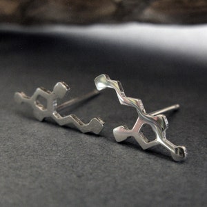 Pode incluir: Um par de brincos de pressão de prata em forma de molécula química. Os brincos são de metal e têm um acabamento brilhante.