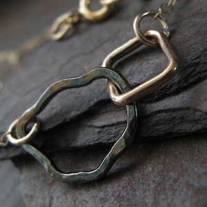 May include: Close-up of a gold-toned necklace featuring a hammered, irregular-shaped link and a square link. The links are connected by small circular rings. The necklace is on a dark, textured surface.