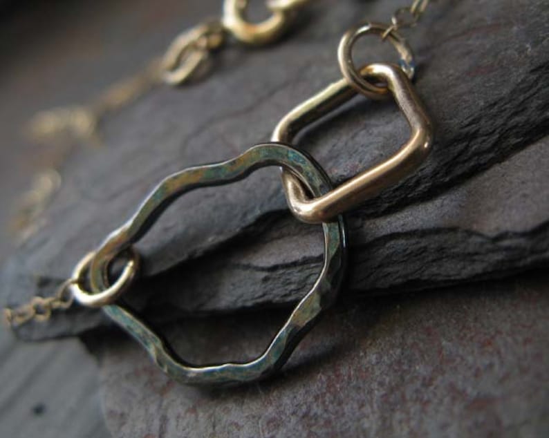 May include: Close-up of a gold-toned necklace featuring a hammered, irregular-shaped link and a square link. The links are connected by small circular rings. The necklace is on a dark, textured surface.