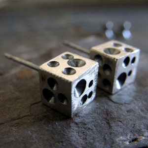 May include: A pair of silver earrings shaped like dice with a textured surface. Each earring has a small post for piercing.