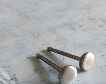 Könnte beinhalten: Ein Paar silberfarbene Ohrstecker mit kleinen, runden, flachen Scheiben und einer gebürsteten Metalloberfläche. Jeder Ohrstecker verfügt über einen geraden Stift, der von der Rückseite des runden Kopfes absteht.