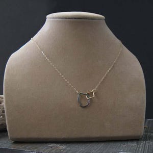 May include: A delicate gold-toned necklace with two geometric pendants: a square and a curved shape. The necklace is displayed on a beige mannequin bust against a dark background. The chain is fine and the pendants are small.