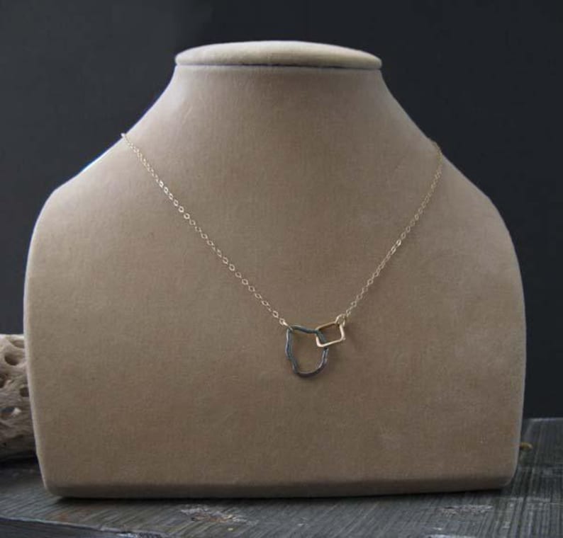 May include: A delicate gold-toned necklace with two geometric pendants: a square and a curved shape. The necklace is displayed on a beige mannequin bust against a dark background. The chain is fine and the pendants are small.