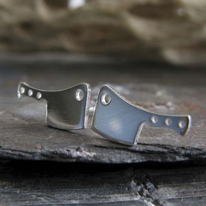 May include: A pair of silver earrings shaped like butcher knives. The earrings are small and delicate, and they have a brushed silver finish.
