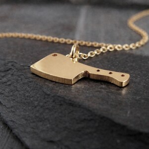 May include: A gold-colored cleaver pendant necklace. The pendant is a miniature cleaver with a handle and a blade. The necklace chain is also gold-colored. The pendant is hanging on a dark surface.
